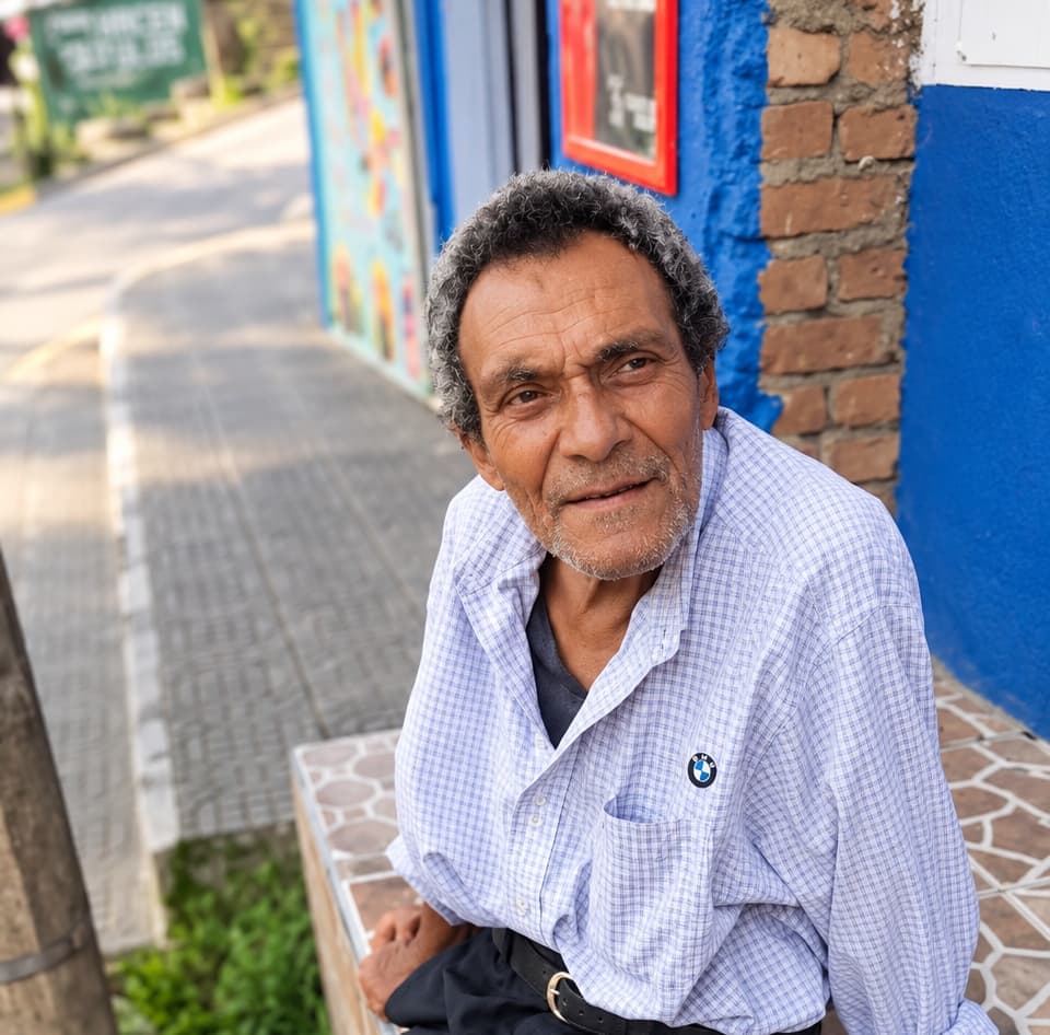 Smiling elderly man in a checked shirt sitting on a bench outside a blue building