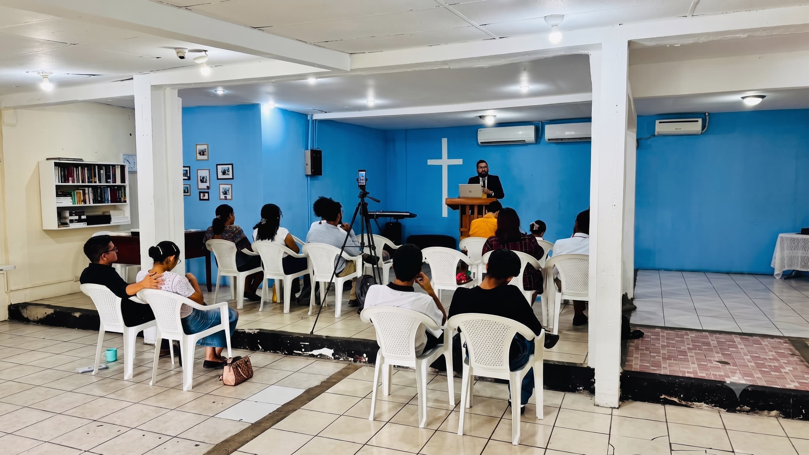 Pastor preaching from a podium to a small congregation in a blue church hall with a white cross