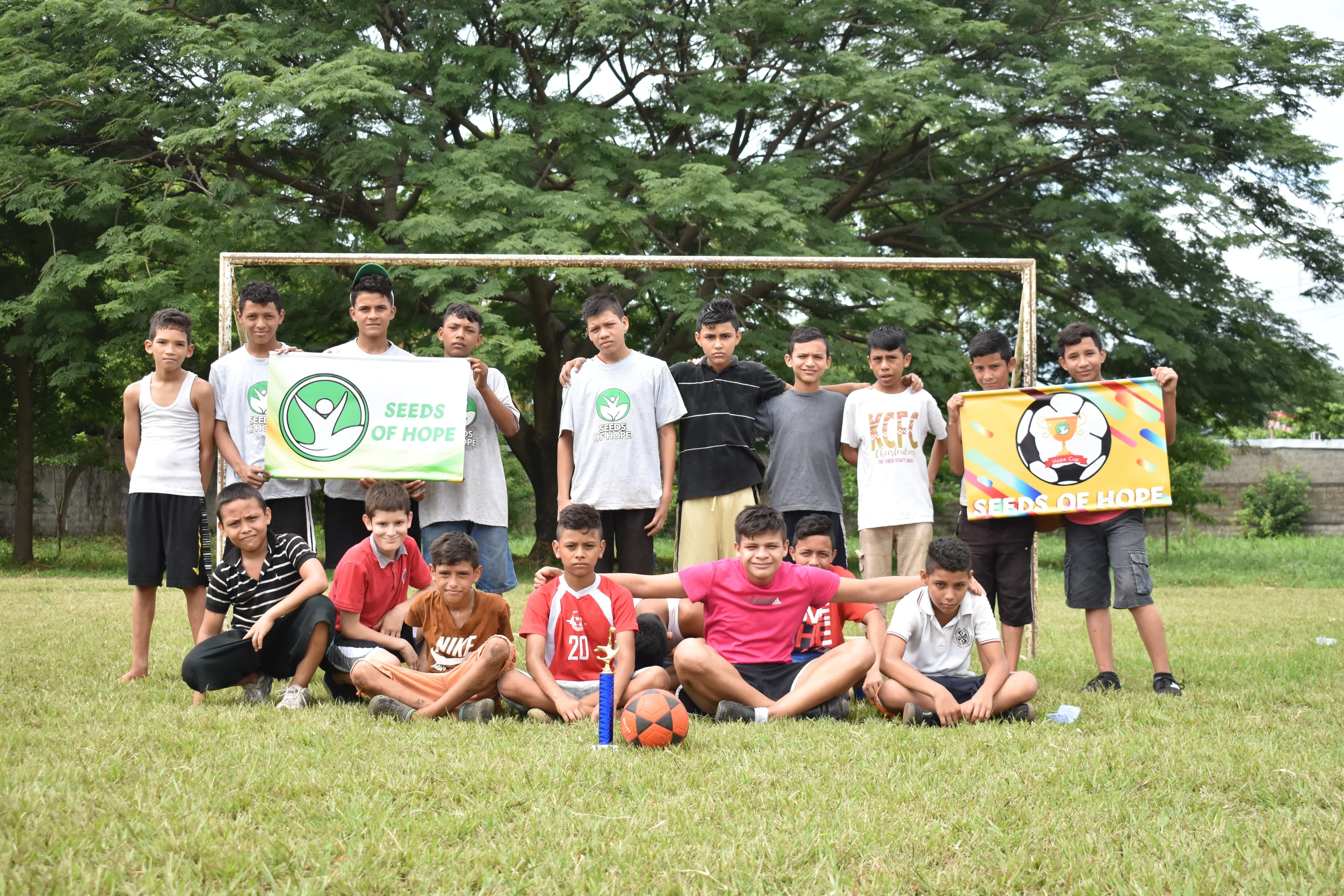 Group of boys posing on a grass field in front of a goal, holding Seeds of Hope banners and a trophy
